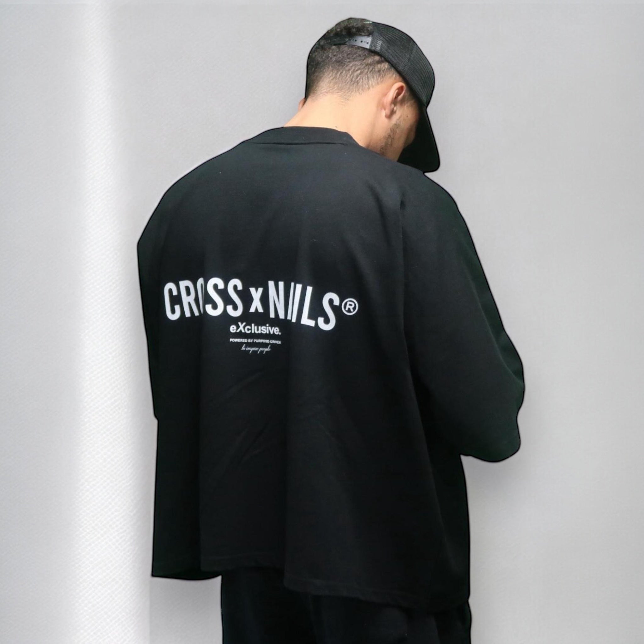 Male model facing away, wearing a black oversized t-shirt with bold white CROSSxNAILS® eXclusive. branding on the back, styled with a black cap, against a neutral grey background.